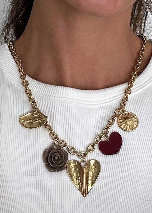 Collier en acier inoxydable doré avec pendentifs décoratifs : cœur rouge, grand cœur doré martelé, rose sculptée marron et lèvres dorées, posé sur une surface claire.