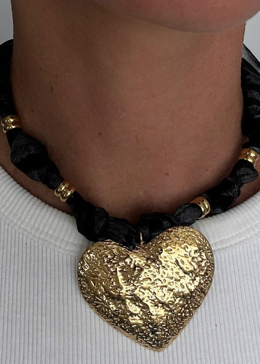Collier ras-de-cou avec ruban noir tressé, perles dorées et grand pendentif cœur martelé doré, porté autour du cou.
