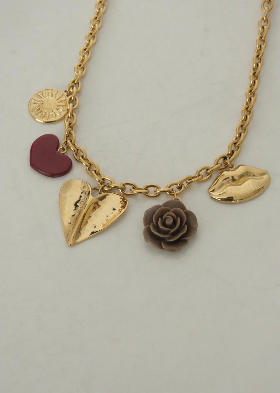 Collier en acier inoxydable doré avec pendentifs décoratifs : cœur rouge, grand cœur doré martelé, rose sculptée marron et lèvres dorées, posé sur une surface claire.