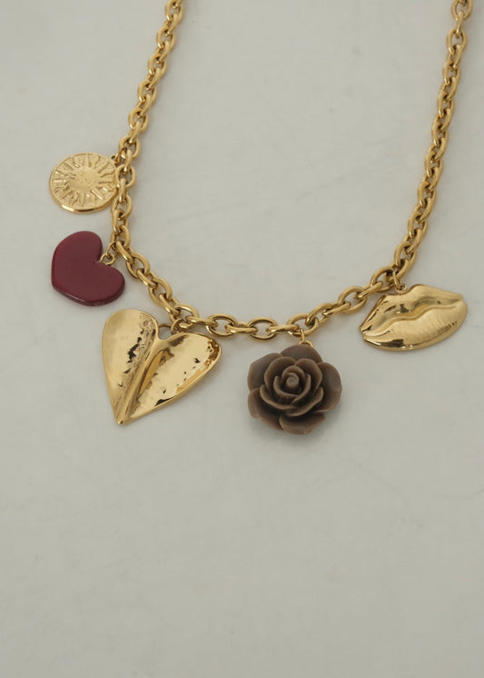Collier en acier inoxydable doré avec pendentifs décoratifs : cœur rouge, grand cœur doré martelé, rose sculptée marron et lèvres dorées, posé sur une surface claire.